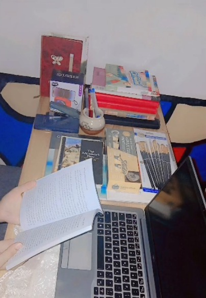 A student's desk: stacked textbooks and notebooks, a pen holder, an open book held in one hand, and a laptop.